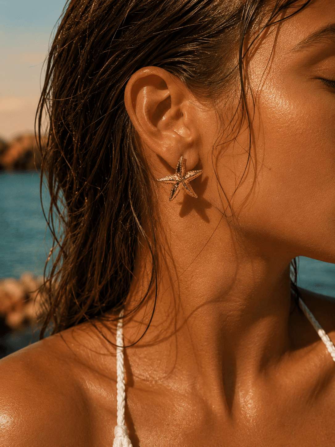 Gros plan macro sur une oreille portant la boucle Astréa au bord de la mer, peau dorée, cheveux mouillés de sel