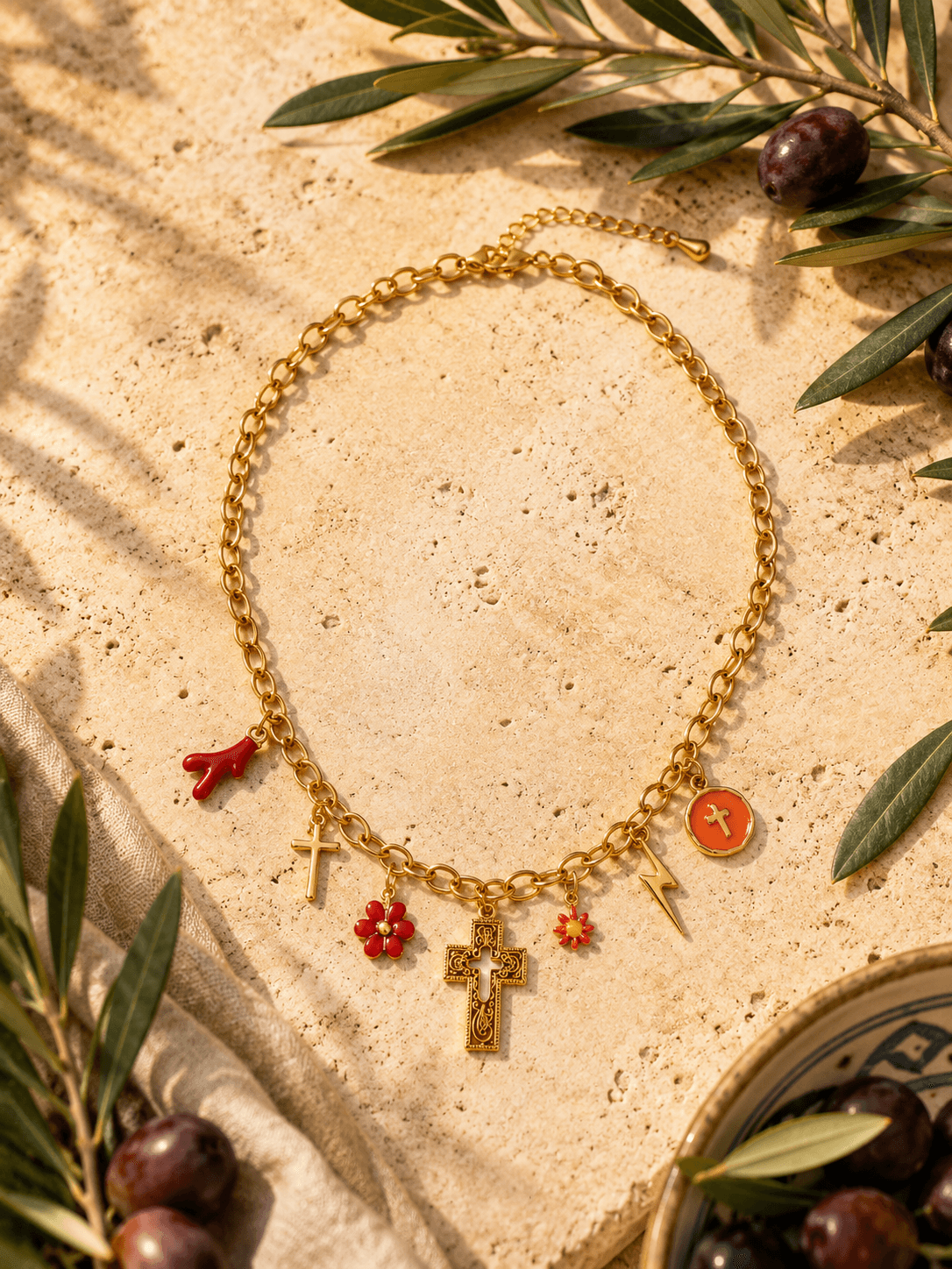 Collier Corallia posé sur un bloc de travertin crème, branches d'olivier avec olives noires, coupe en céramique bleue et blanche style Moustiers, ombre dansante de feuillage
