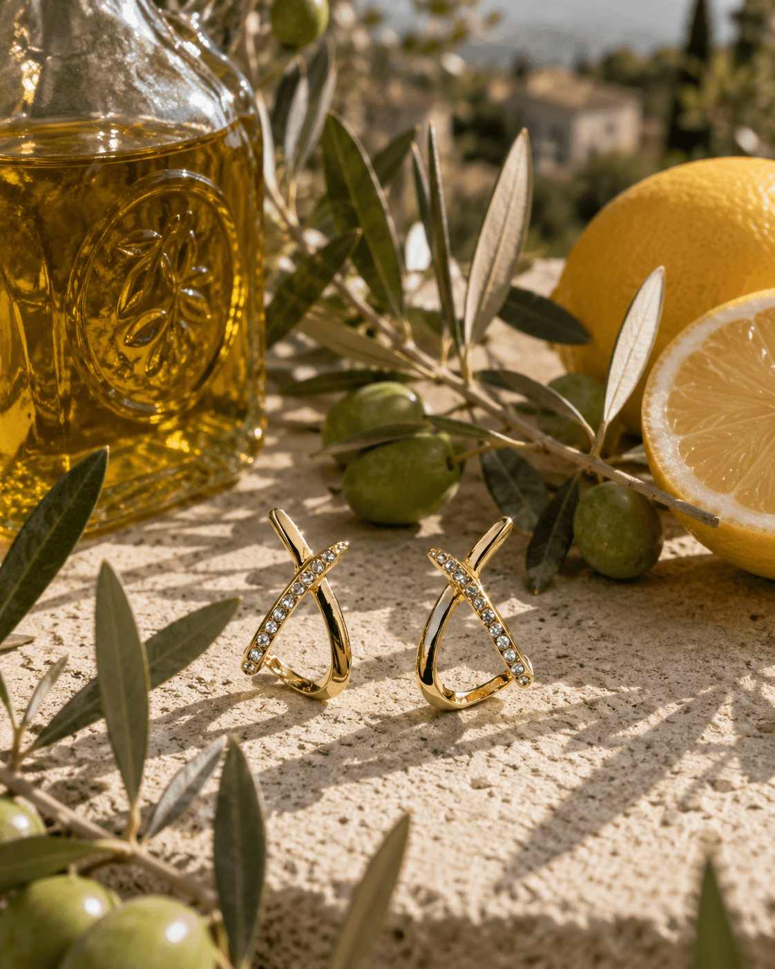 Boucles Croisette posées sur un bloc de travertin, bouteille d'huile d'olive gravée et citron entier en arrière-plan, branches d'olivier avec olives vertes, village provençal flou à l'horizon