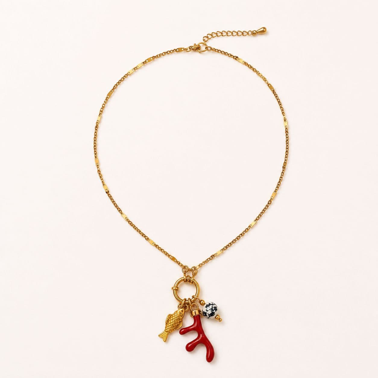 Collier Egea à plat sur fond crème pâle, chaîne fine à maillons alternés barre rectangulaire et maillon rond, cluster pendentif au bas — petit poisson d'or, brin de corail rouge méditerranéen, perle de jaspe dalmatien noir-et-blanc