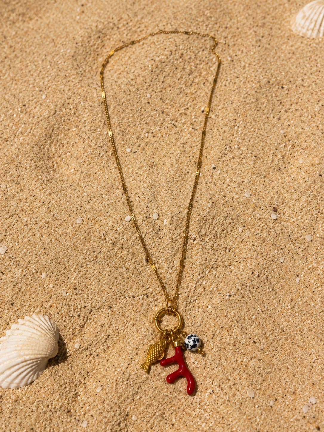 Collier Egea posé sur le sable doré, chaîne fine étirée en demi-cercle, cluster pendentif en bas et coquillage Saint-Jacques nacré à gauche, lumière du soleil zénithal