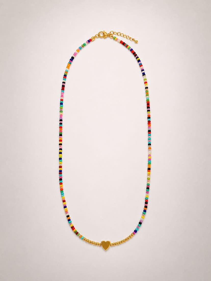 Collier Festa à plat sur fond crème, fil de perles de rocaille multicolores enfilées (rose, rouge, bleu, vert, turquoise, jaune, orange, noir, blanc), petit cœur d'or au centre encadré de six perles dorées rondes
