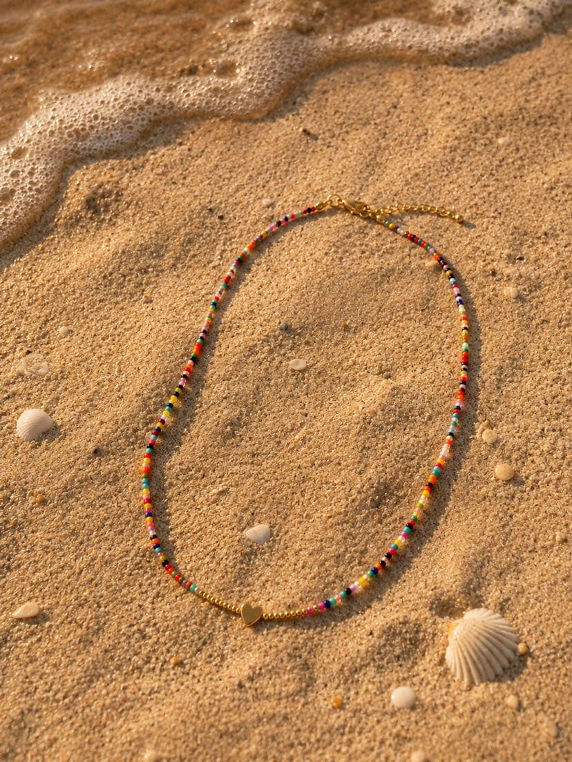 Collier Festa posé sur le sable doré, étiré en demi-cercle, coquillages Saint-Jacques crème éparpillés autour, vague d'écume au loin, ombre de fin d'après-midi