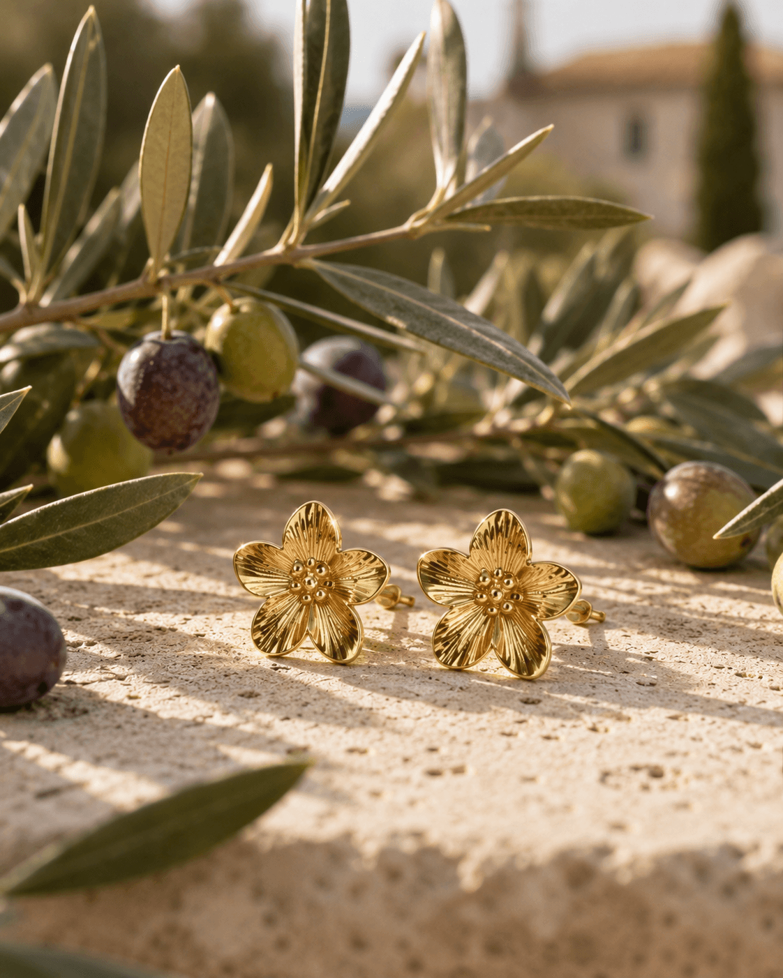Boucles Floralia posées sur un bloc de travertin crème, branche d'olivier avec olives vertes et noires en arrière-plan, village méditerranéen flou à l'horizon, lumière dorée de fin d'après-midi