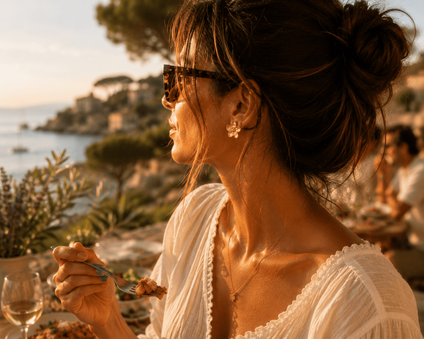 Profil féminin au coucher du soleil, lunettes en écaille, peau dorée, boucle Floralia portée, dîner sur une terrasse en bord de mer méditerranéenne, voiliers à l'ancre