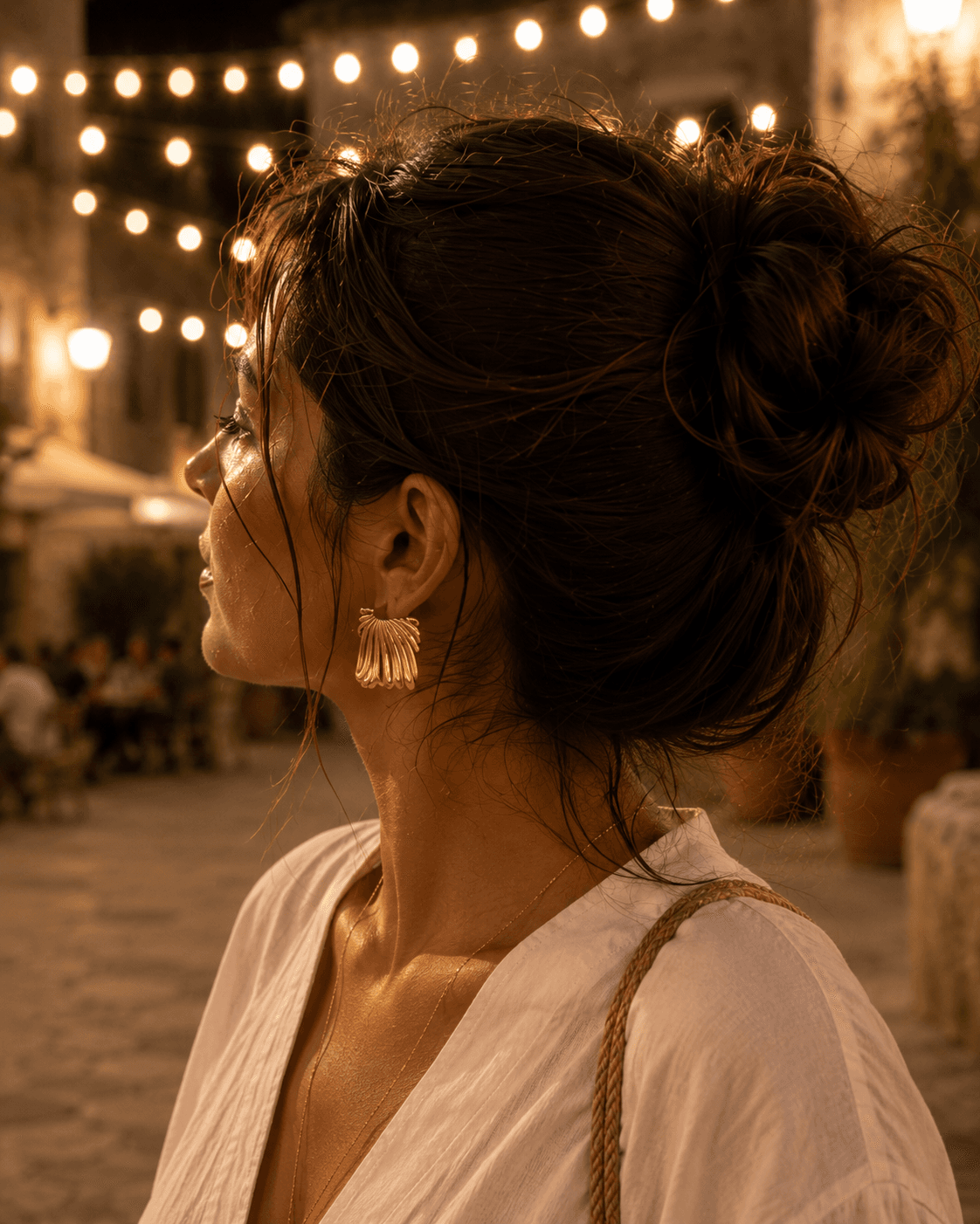 Profil féminin de soir, chignon flou et mèches au vent, boucle Hélios à l'oreille, guirlandes lumineuses au-dessus d'une piazza italienne en pierre