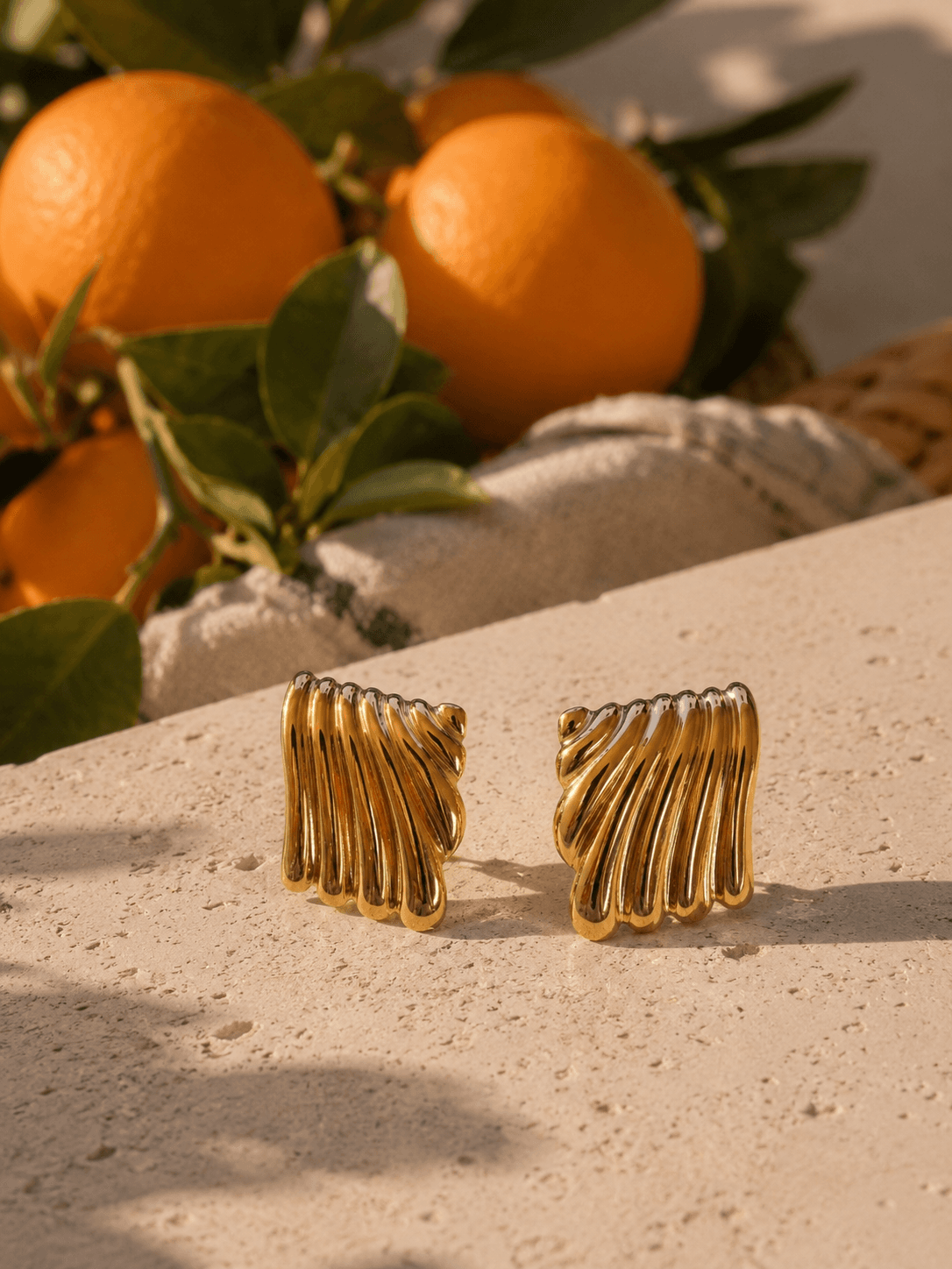 Boucles Hélios posées sur un muret de travertin crème, oranges et feuillage en arrière-plan, lumière franche de midi