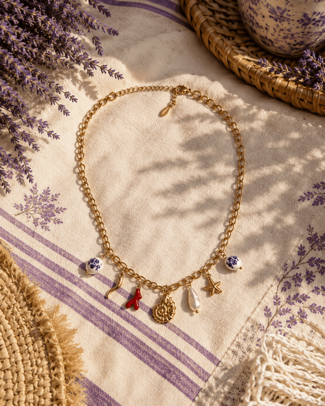 Collier Lavanda posé sur un torchon de lin écru rayé de lavande, brins de lavande à gauche, panier en paille et coupe en céramique de Moustiers à droite, ombre de feuillage sur la table