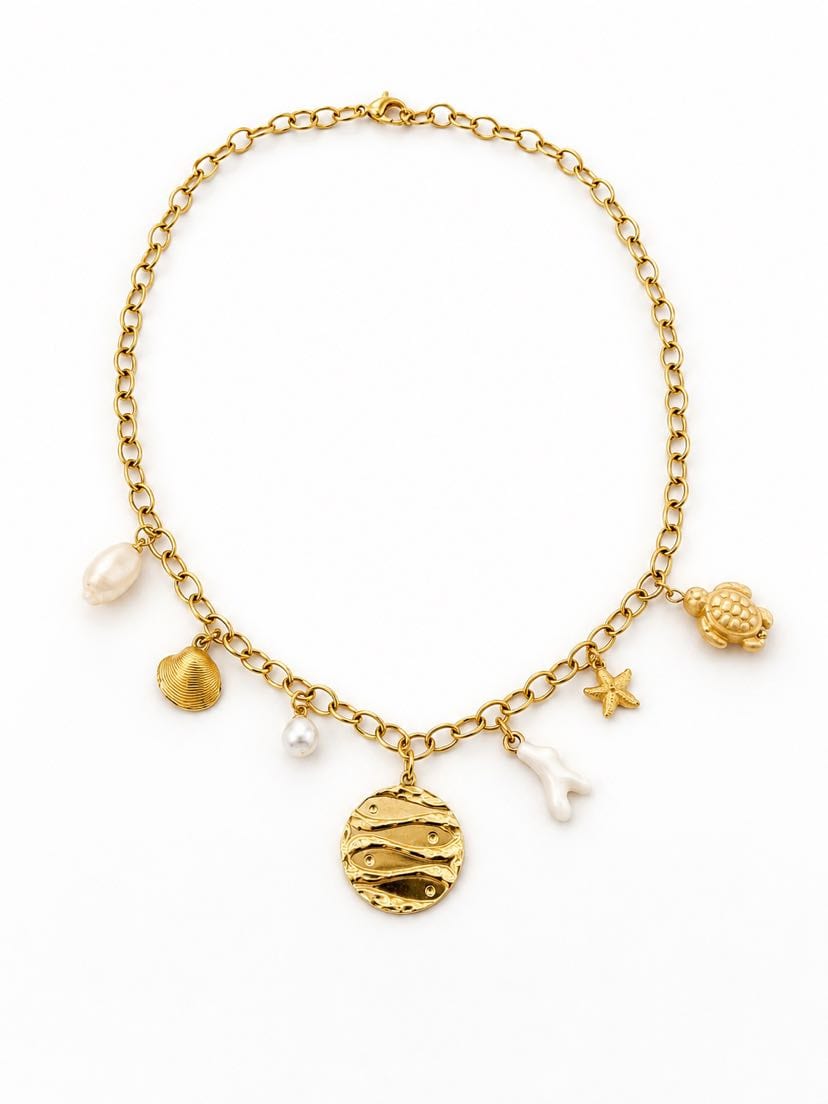 Collier Onda à plat sur fond crème, chaîne forçat épaisse d'or et 7 pendentifs : médaillon ondulé ciselé au centre, perle baroque, coquillage bombé, perle ronde, brin de corail blanc, étoile de mer, petite tortue d'or