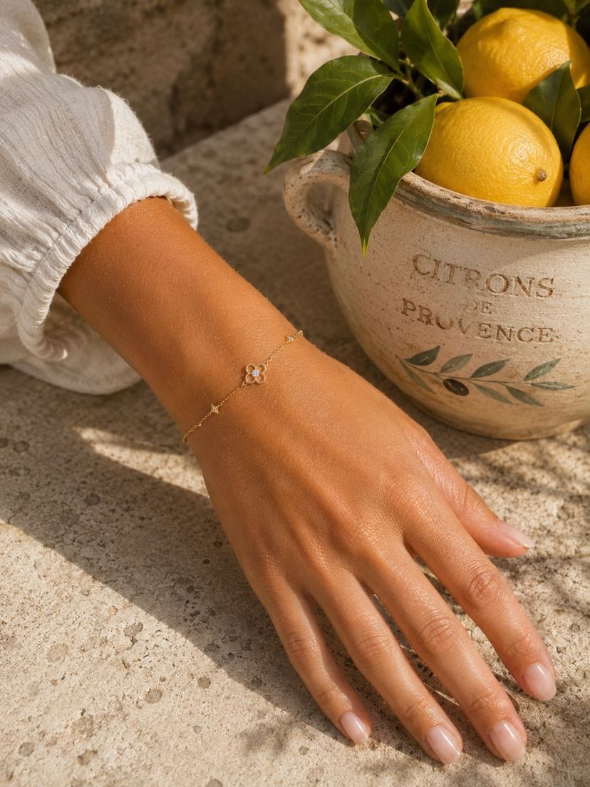 Main bronzée posée à plat sur du travertin clair en Provence, manche blanche tunisienne en mousseline froissée, bracelet Cicadu chaîne fine or jaune avec trèfle 4 pétales central et deux étoiles latérales pavées au poignet, jarre en céramique 'Citrons de Provence' avec citrons jaunes et feuillage d'olivier à droite