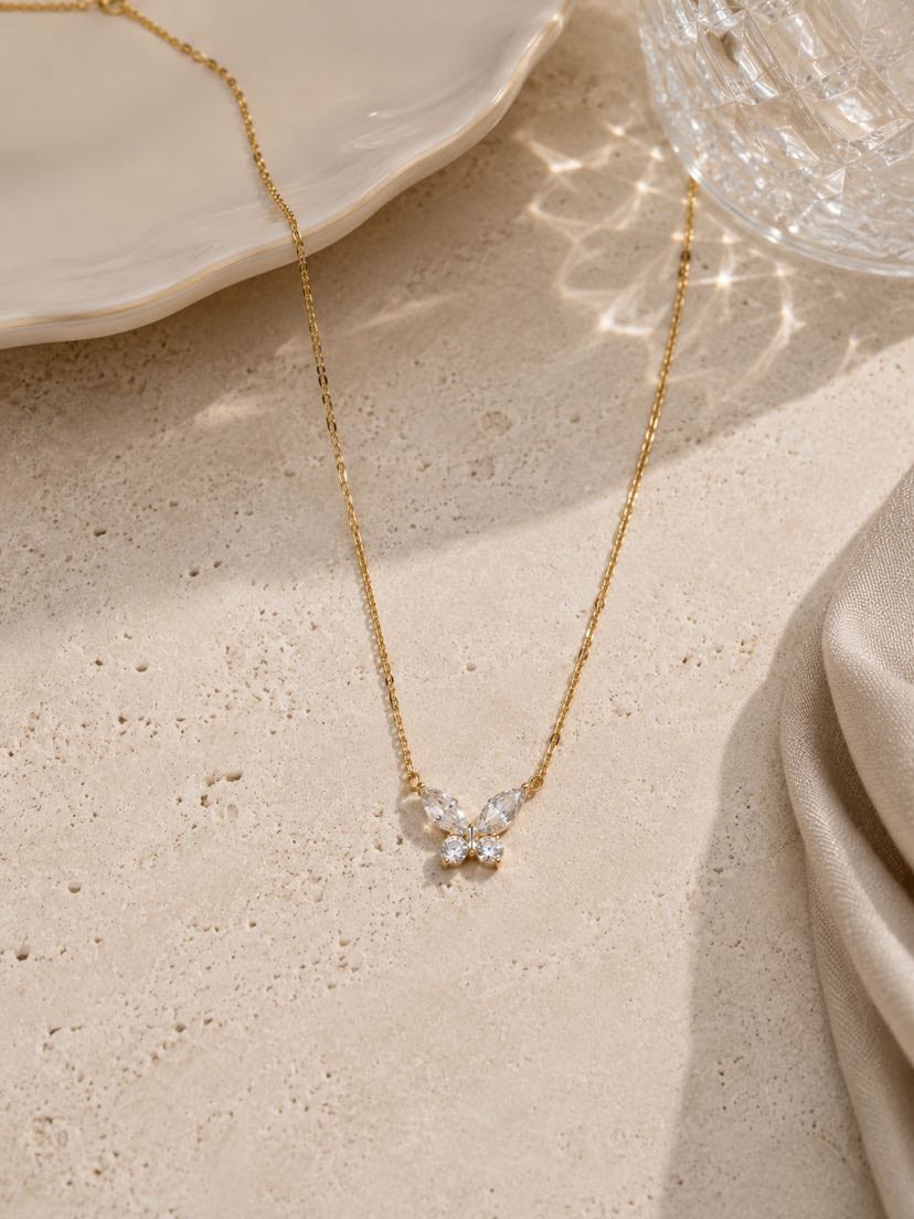 Collier Sigillo en packshot vu de dessus, chaîne fine dorée traversant la composition, médaillon rond en émail noir cerclé d'or au centre, posé sur travertin crème à côté d'une assiette en porcelaine ondulée et d'un verre en cristal taillé qui projette des reflets en éventail