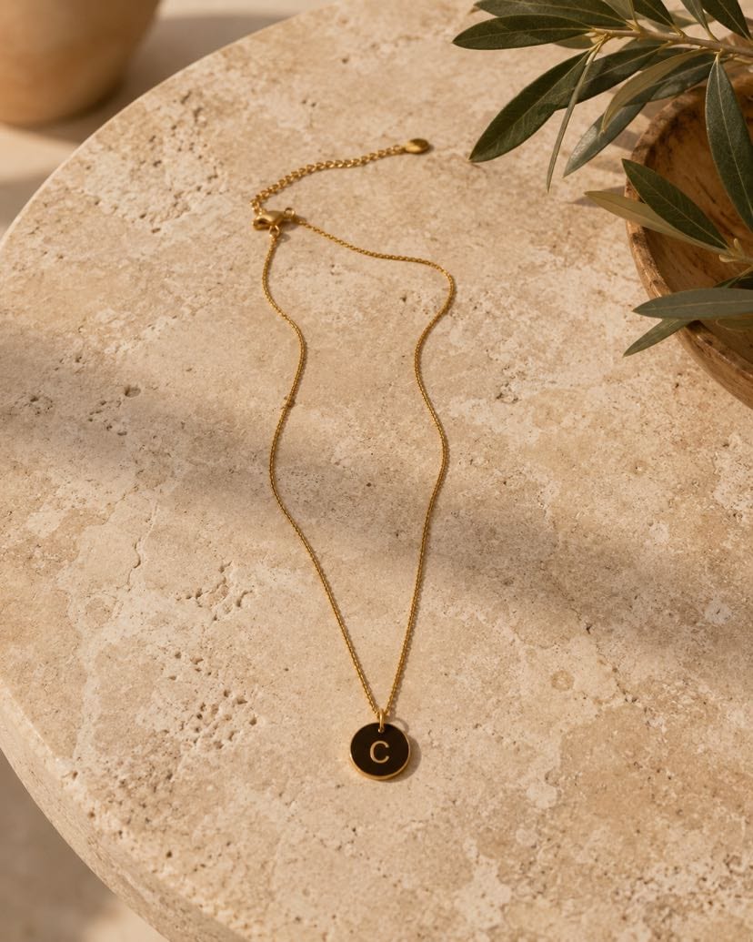 Collier Sigillo posé en serpentin sur une table ronde en travertin crème, chaîne fine dorée qui ondule avec sa chaînette de rallonge et plaque ovale signature visibles en haut, médaillon rond en émail noir gravé lettre C dorée centré en bas, jarre artisanale en céramique terracotta avec olivier à droite, ombre tachetée d'olivier sur la pierre, lumière dorée d'après-midi