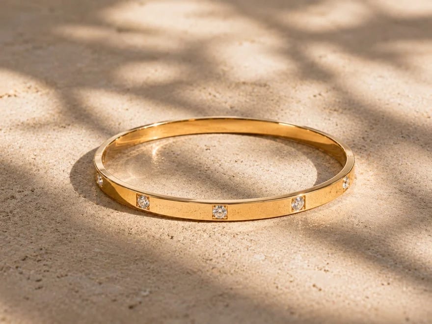 Bracelet jonc Tramonto plat or jaune posé sur du travertin sablé doré, zircons sertis flush captant la lumière, ombres mouchetées de feuillage projetées sur la pierre, reflet doré du jonc sur le sable, lumière dorée d'après-midi