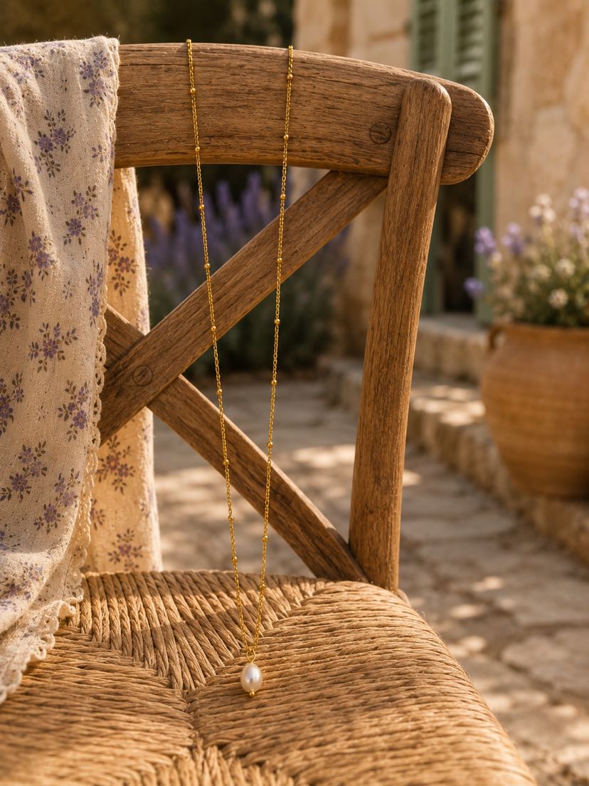 Collier Perla déposé sur une chaise en bois ancien à assise en paille tressée, robe imprimée fleurs pastel suspendue à côté, lavandes en pot terracotta floues en arrière-plan, lumière de midi