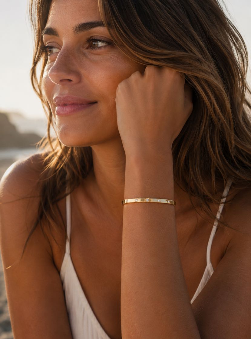 Portrait brune cheveux longs ondulés au coucher du soleil sur la plage, joue posée sur la main fermée, bracelet jonc Tramonto plat or jaune avec petits zircons sertis flush au poignet, sourire doux, falaise calcaire en arrière-plan flou, lumière dorée rasante