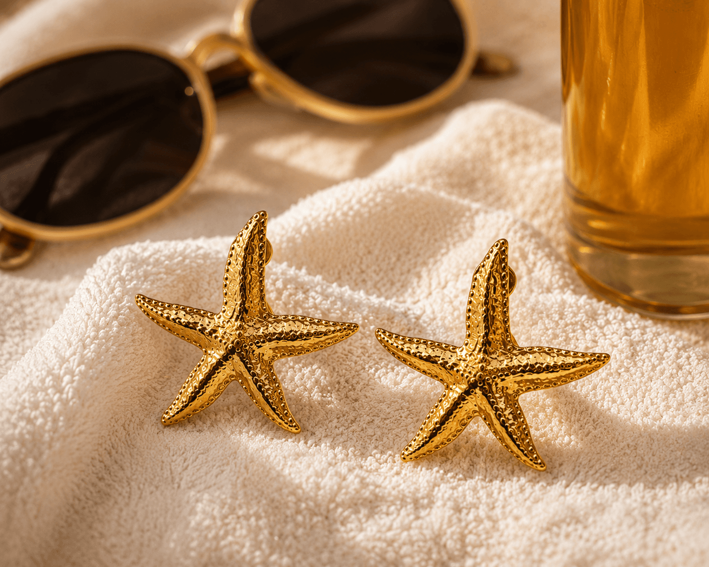 Boucles Astréa posées sur une serviette de plage en éponge écrue, lunettes dorées et verre de limonade dorée à proximité, lumière de fin d'après-midi