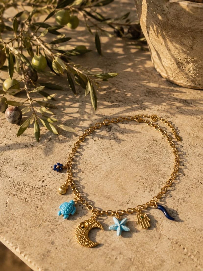 Collier Azulina posé sur une table en travertin doré, chaîne d'or et 7 breloques bleues visibles — tortue et étoile de mer turquoise, croissant de lune ciselé, main de Fatma, fleur de lapis, coquille dorée, cornicello bleu — branche d'olivier avec olives vertes et jarre en terracotta en arrière-plan, ombres mouchetées du soleil, lumière dorée d'après-midi