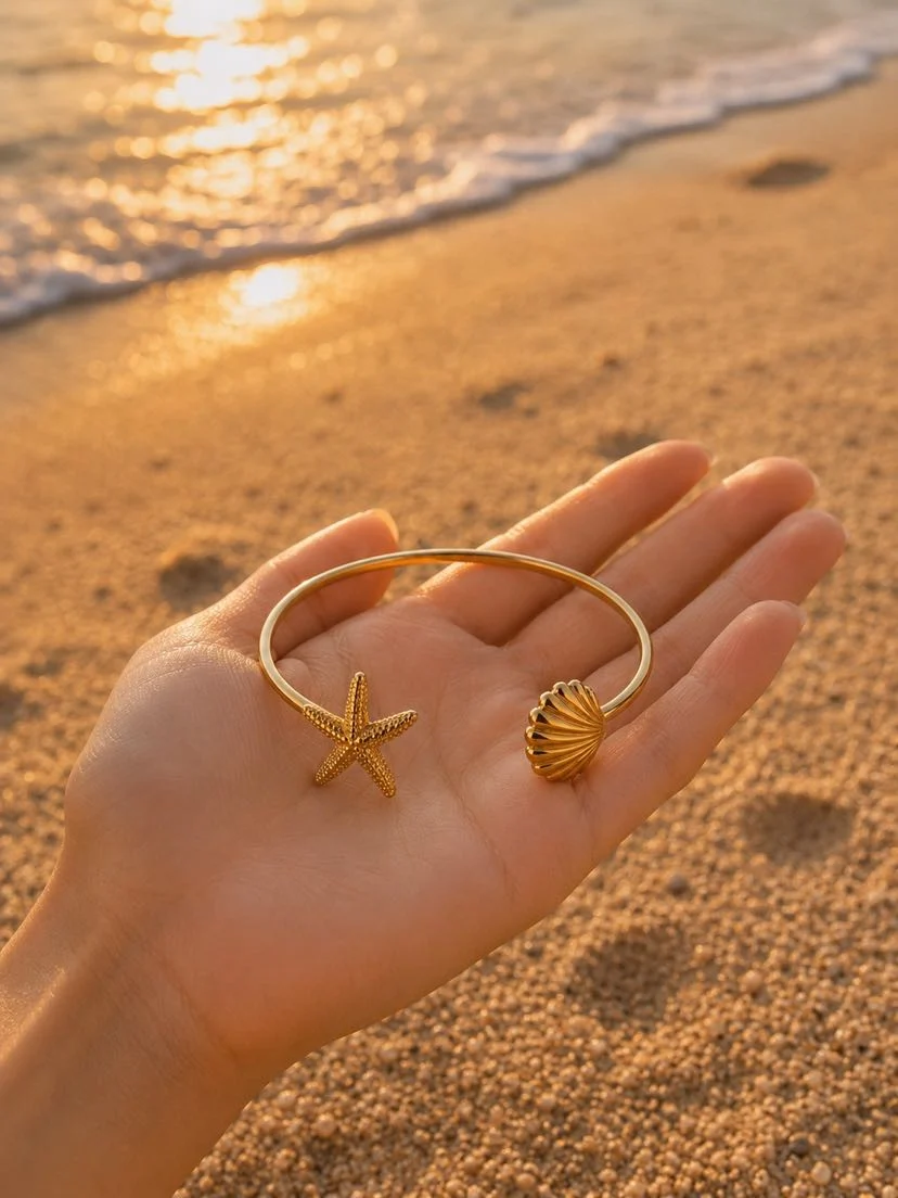 Bracelet jonc Catalina ouvert en plaqué or présenté dans le creux d'une main, étoile de mer ciselée d'un côté et coquille Saint-Jacques bombée de l'autre, sable doré et écume de mer au coucher du soleil en arrière-plan, lumière dorée rasante