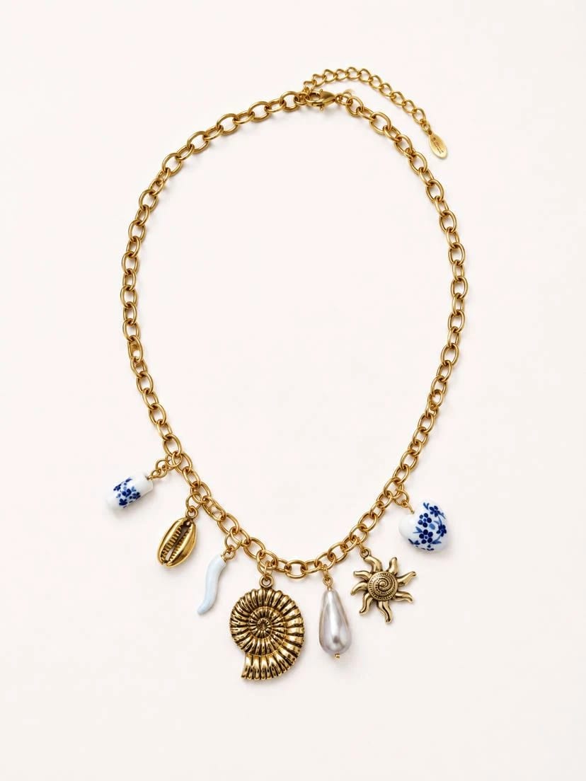 Collier Sol de Oliva à plat sur fond crème, chaîne forçat épaisse d'or et 7 breloques — perle de faïence bleue, coquille cauri dorée, cornicello blanc, ammonite spiralée ciselée centrale, perle baroque blanche en goutte, soleil rayonnant doré, perle de faïence bleue en cœur, fermoir et rallonge visibles en haut