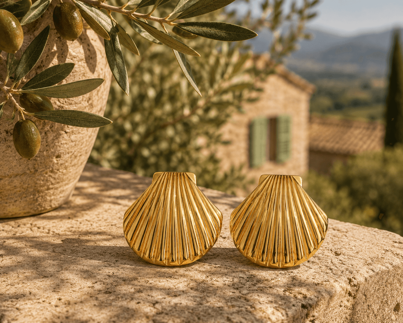 Boucles Vénus posées sur un muret de pierre, jarre en terracotta avec branche d'olivier et olives vertes à gauche, bastide provençale aux volets vert pastel et collines en arrière-plan